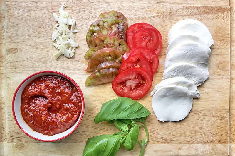 Horizontal top-down image of slices of tomatoes, mozzarella, garlic, basil, and a bowl of marinara on a wooden cutting board.