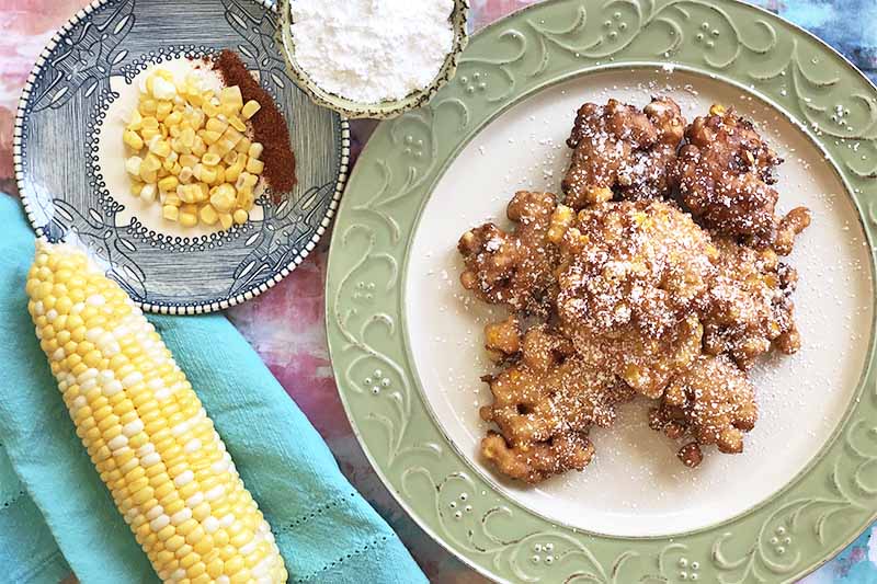 Horizontal image of deep-fried morsels on a green-trimmed plate next to corn and powdered sugar on a blue towel.
