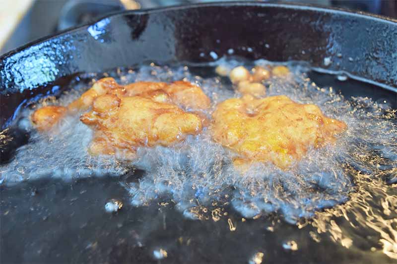 Horizontal image of two golden-brown morsels being fried in oil in a black skillet.