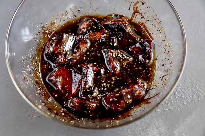 Horizontal image of a glass bowl with marinated meat pieces in a dark brown liquid.