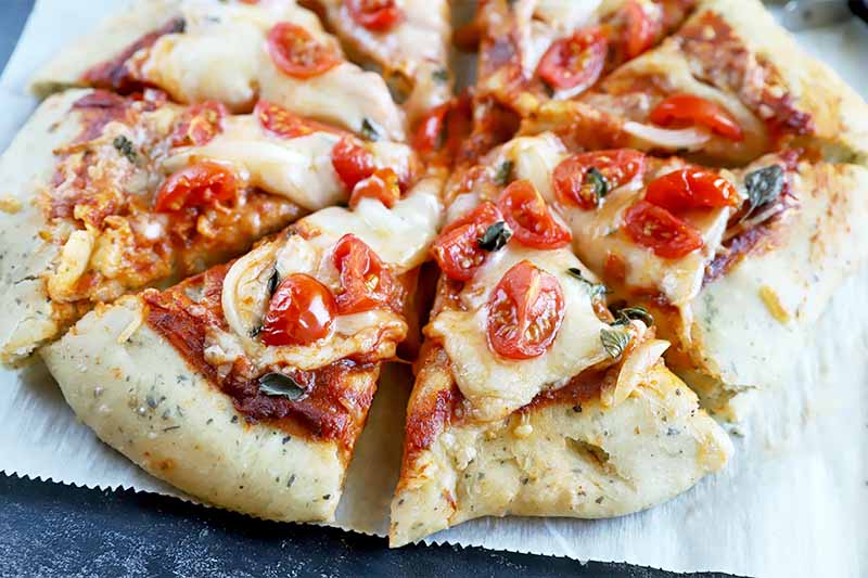 Horizontal image of a sliced pizza topped with tomatoes and melted cheese on a parchment paper.