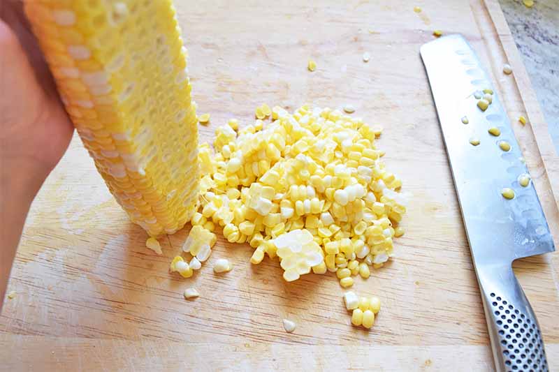Horizontal image of removing kernels from a cob with a metal knife on a wooden cutting board.