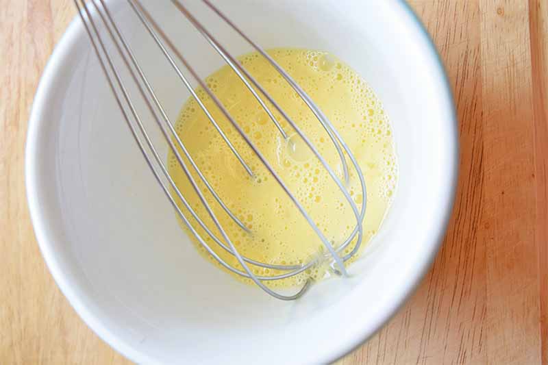 Horizontal image of a whisk mixing a yellow liquid in a white bowl on a wooden surface.