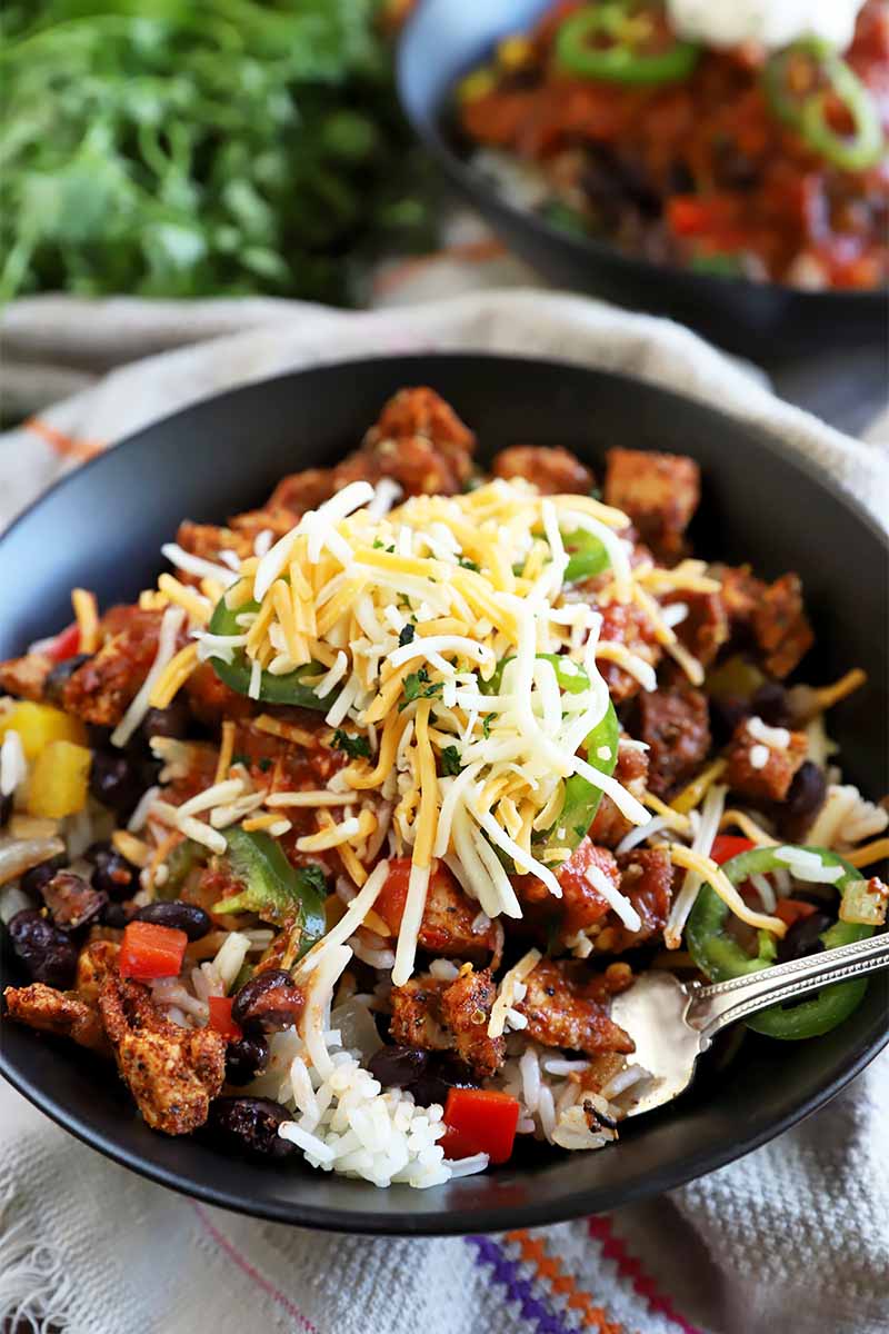 Vertical mage of a black bowl with a white rice base topped with spiced meat, vegetables, beans, jalapeno slices, and cheddar cheese, with a fork inserted into a part of the dish, with another bowl and cilantro in the background.