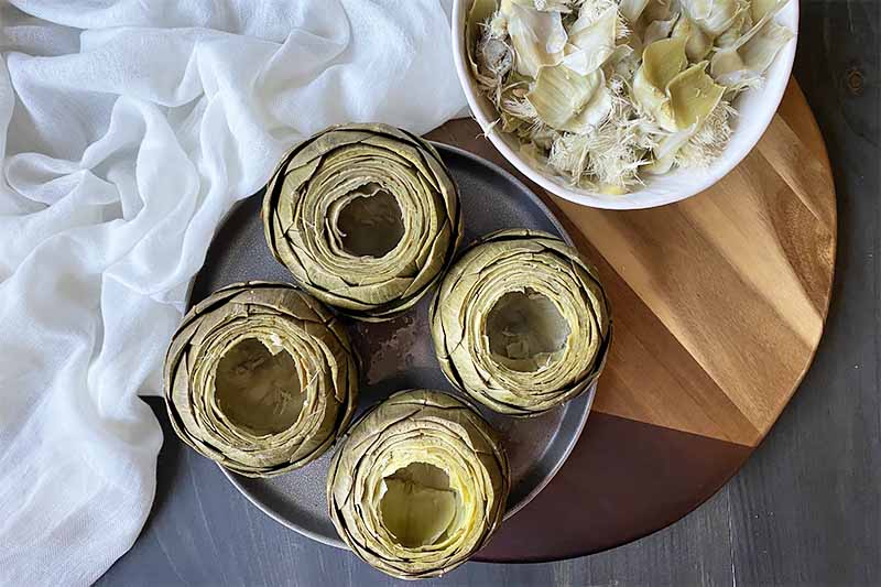Horizontal image of four hollowed out boiled artichokes on a plate next to a bowl of their interior filling on a wooden cutting board.