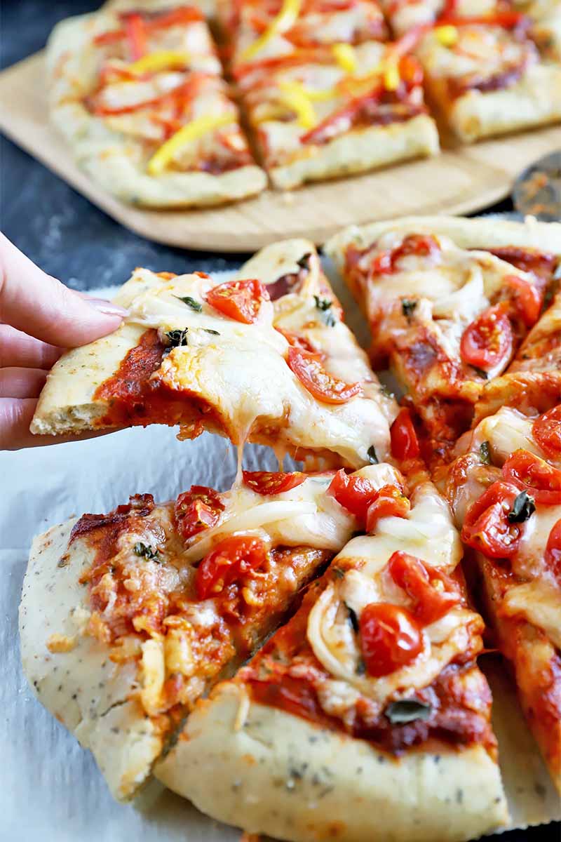 Vertical image of a hand picking up a slice of pizza with gooey cheese on top of parchment paper, in front of the same recipe on a wooden cutting board.