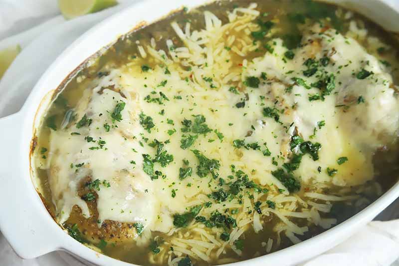 Horizontal image of a white casserole dish filled with with meat, sauce, melted cheese, and fresh herbs.
