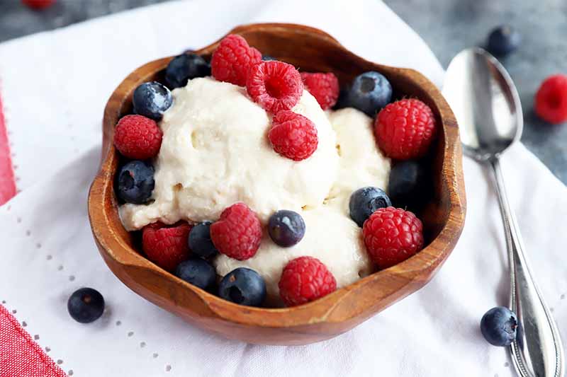 Horizontal image of a wooden bowl filled with scoops of vanilla ice cream topped with raspberries and blueberries on a white napkin next to a metal spoon.