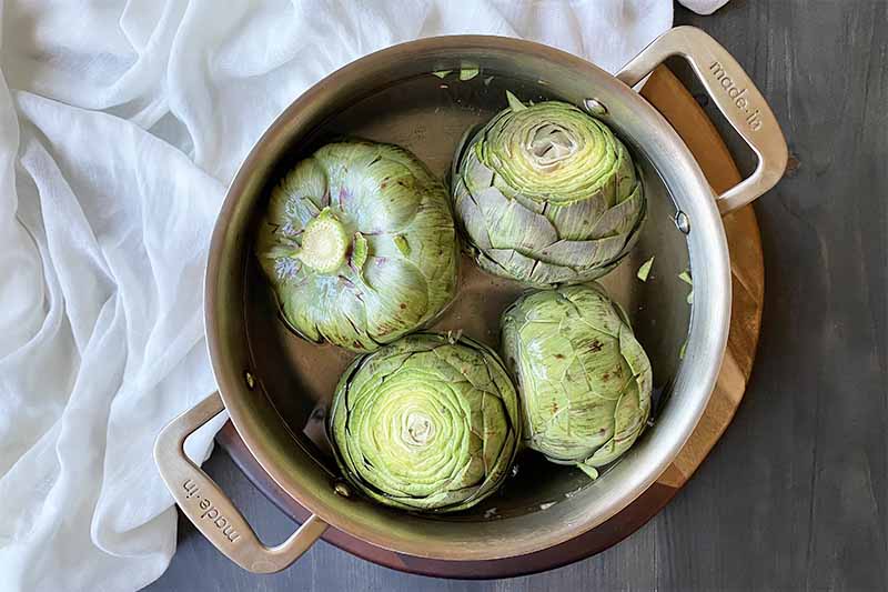 Horizontal image of a pot with water and four whole artichokes.
