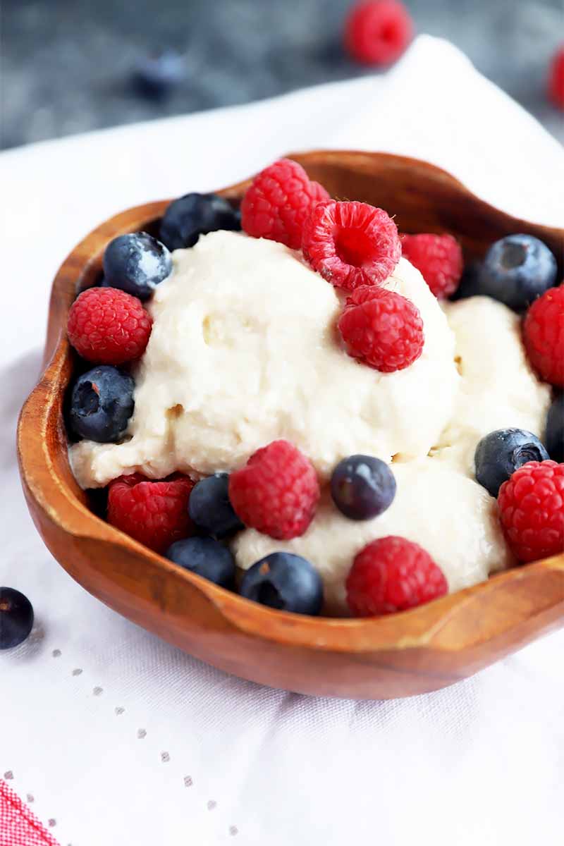 Vertical image of a wooden bowl with scoops of vanilla ice cream topped with raspberries and blueberries on a white napkin on a dark table.