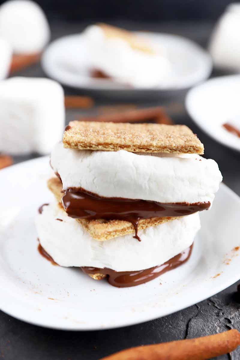Vertical image of a stack of two s'mores with fluffy marshmallow and melted chocolate on a white plate on a gray surface.