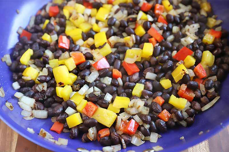 Horizontal image of a blue pan cooking an assortment of mixed chopped vegetables and black beans.