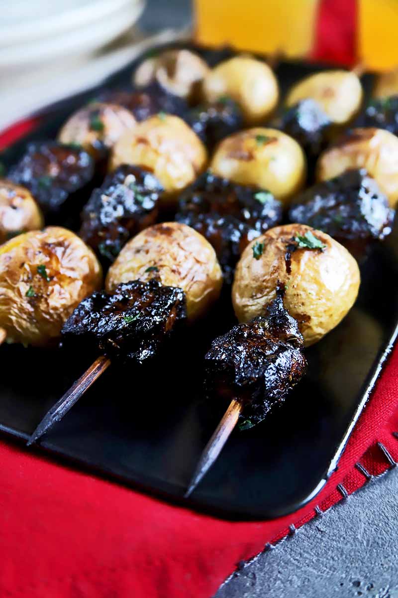 Vertical close-up image of skewers of grilled meat and spuds on a black plate on a red towel