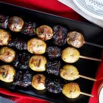 Horizontal image of four grilled meat and potato skewers on a black rectangular plate on a red towel next to two glasses of beer.
