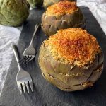 Horizontal image of two whole artichokes with breadcrumbs filling on a dark slate on a towel next to forks.