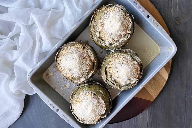 Horizontal image of four artichokes topped with grated cheese in a metal pan.