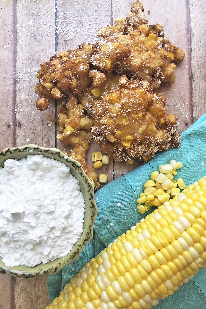 Vertical top-down image of a pile of deep-fried morsels next to a bowl of powdered sugar, a blue towel, and a whole corn on the cob.