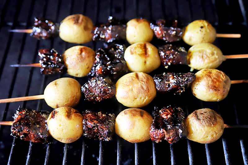 Horizontal image of a grill with four skewers of alternating potato and beef pieces.