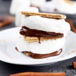 Horizontal image of a stack of two s'mores with fluffy marshmallow and melted chocolate on a white plate on a gray surface.