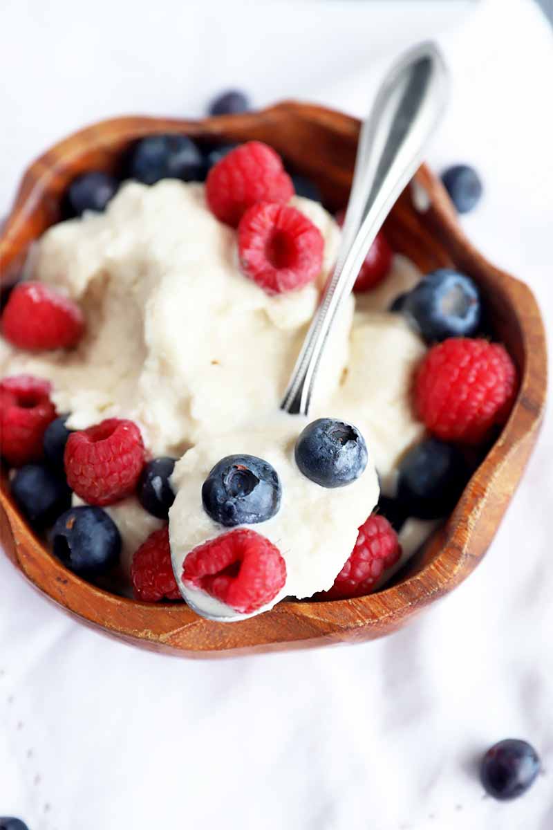 Vertical image of a wooden bowl on a white napkin filled with vanilla ice cream and berries, and a spoon holding some of the dessert.