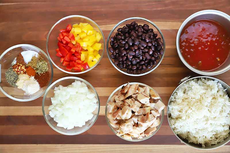 Horizontal image of a wooden table with bowls of assorted prepped vegetables, black beans, chicken, and salsa.