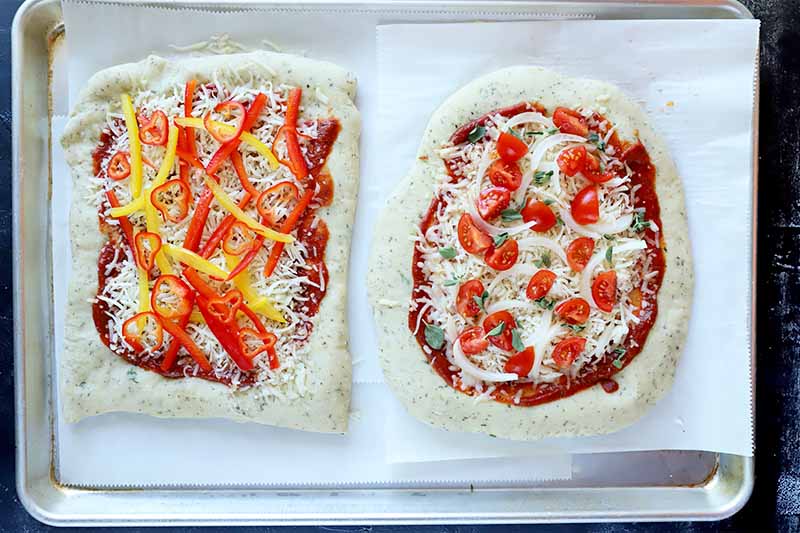 Horizontal image of two unbaked crusts on parchment paper topped with tomato sauce, cheese, and an assortment of vegetables.