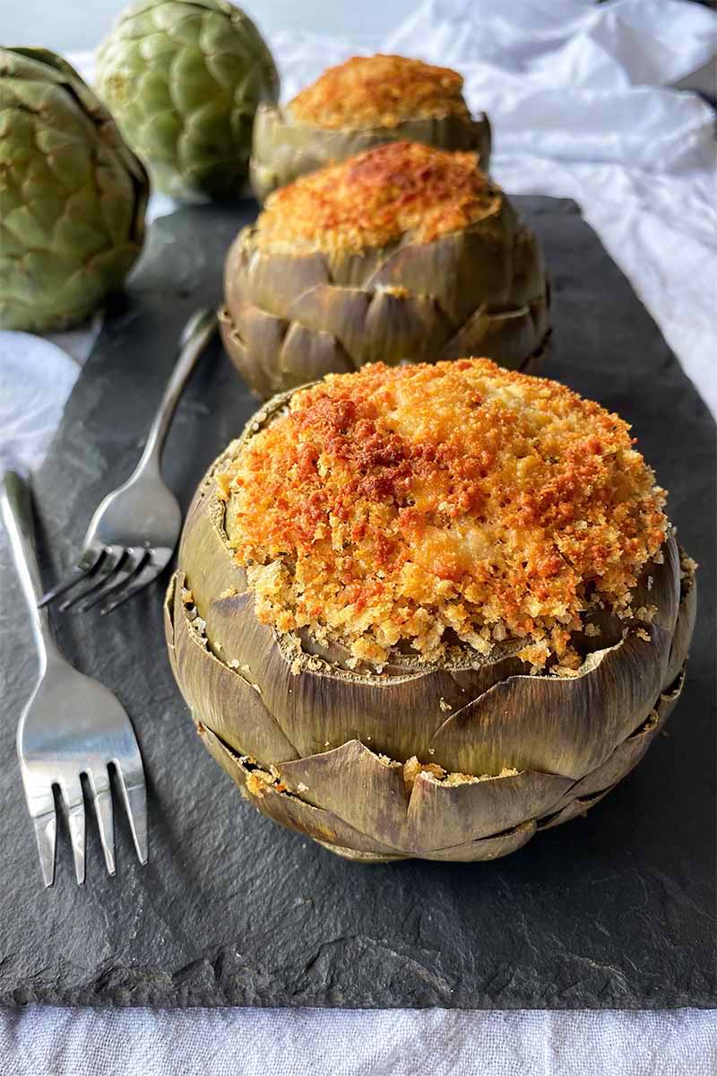 Vertical image of three stuffed artichokes on a slate next to two metal forks.