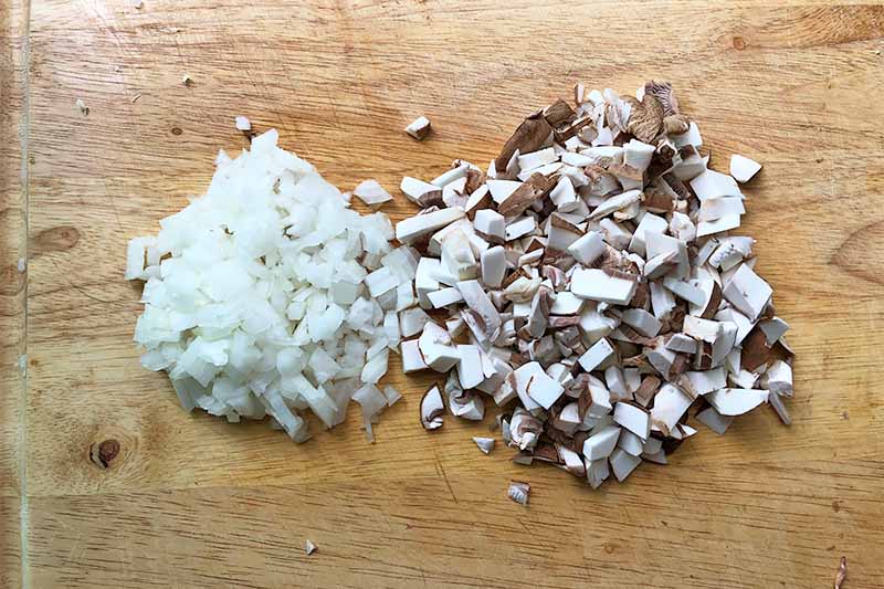 Horizontal image of finely chopped onions and mushrooms on a wooden cutting board.