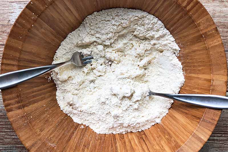 Horizontal image of mixing together coconut oil and dry ingredients with two forks in a wooden bowl.