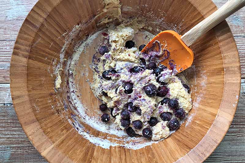 Horizontal image of a wet, thick dough with blueberries in a wooden bowl being stirred by an orange spatula.