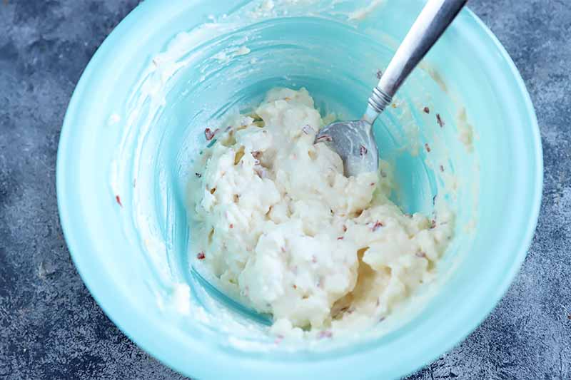 Horizontal image of a blue bowl filled with a seasoned mayonnaise spread mixed by a metal spoon.
