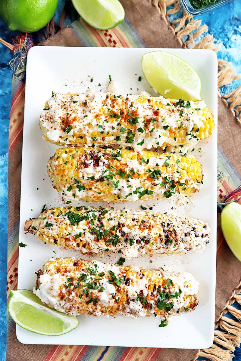 Vertical top-down image of a row of Mexican street corn on a rectangular white plate next to whole and sliced limes.