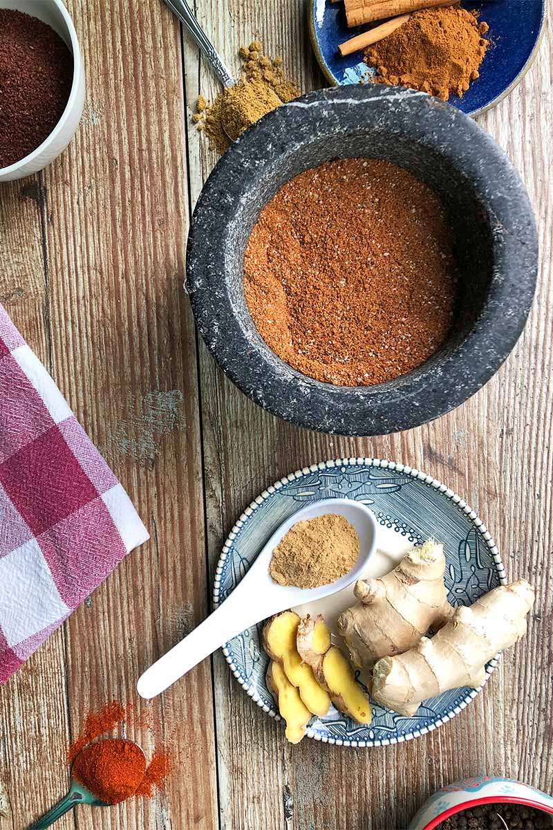 Vertical top-down image of a gray mortar filled with a dark orange spice mixture on a wooden table next to a plate with fresh and ground ginger in a spoon, and other metal spoons with ground spices next to a checkered towel.