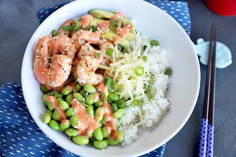 Horizontal image of a white bowl with rice, edamame, and shrimp on a blue napkin next to chopsticks and a red cup.
