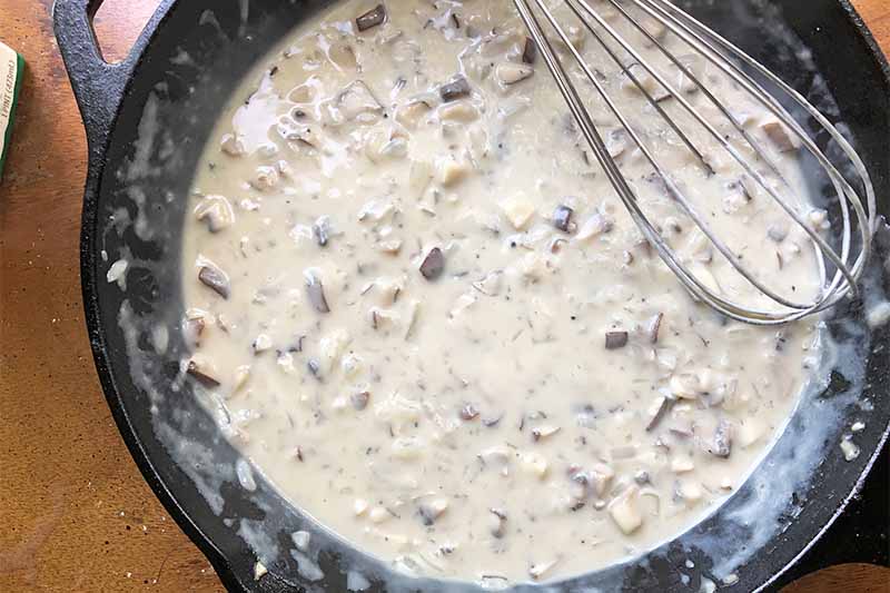 Horizontal image of a cast iron skillet filled with a creamy liquid and chopped brown vegetables with a metal whisk on the side.