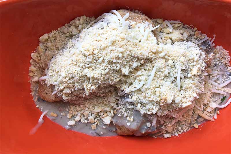 Horizontal image of a red baking dish filled with two raw poultry breasts covered in a thick cream, breadcrumbs, and shredded cheese.
