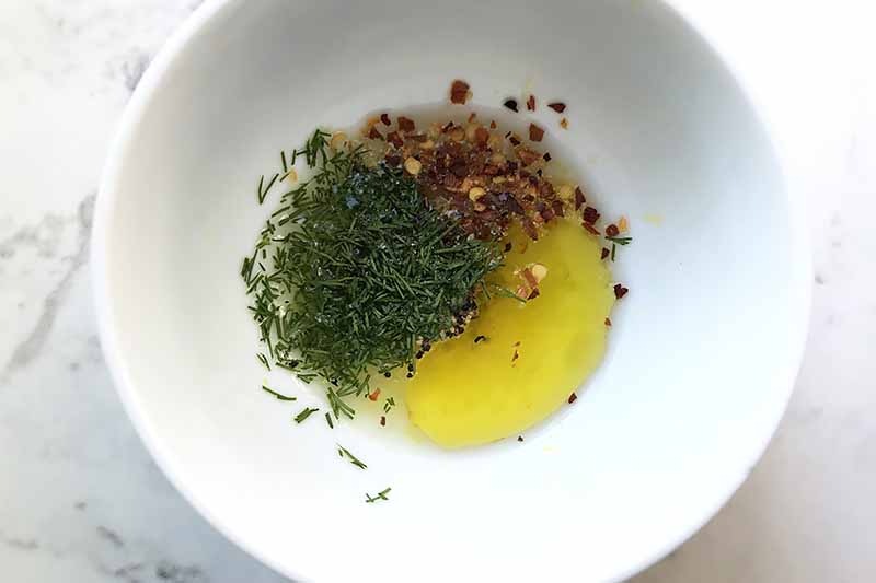 Horizontal image of a white bowl with dill, red pepper flakes, and oil.