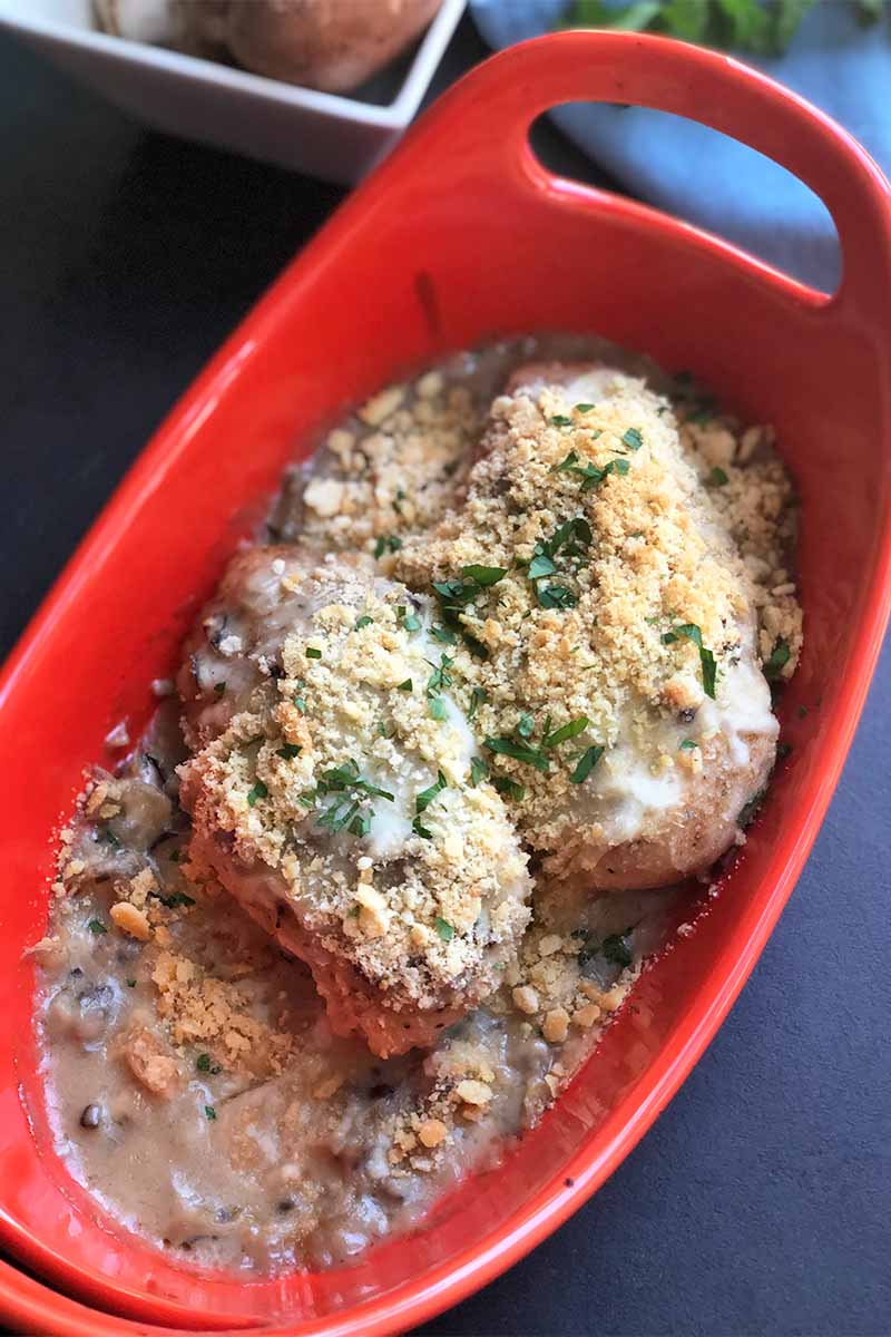 Vertical image of a red baking dish with two poultry breasts topped with sauce, breadcrumbs, and chopped fresh herbs.