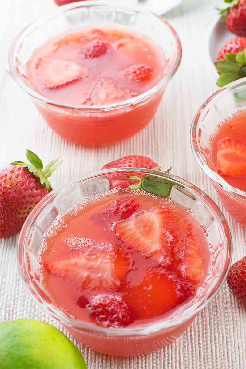 Vertical image of three small glass bowls filled with a clear strawberry dessert with sliced fruit on a light surface next to fresh fruit.