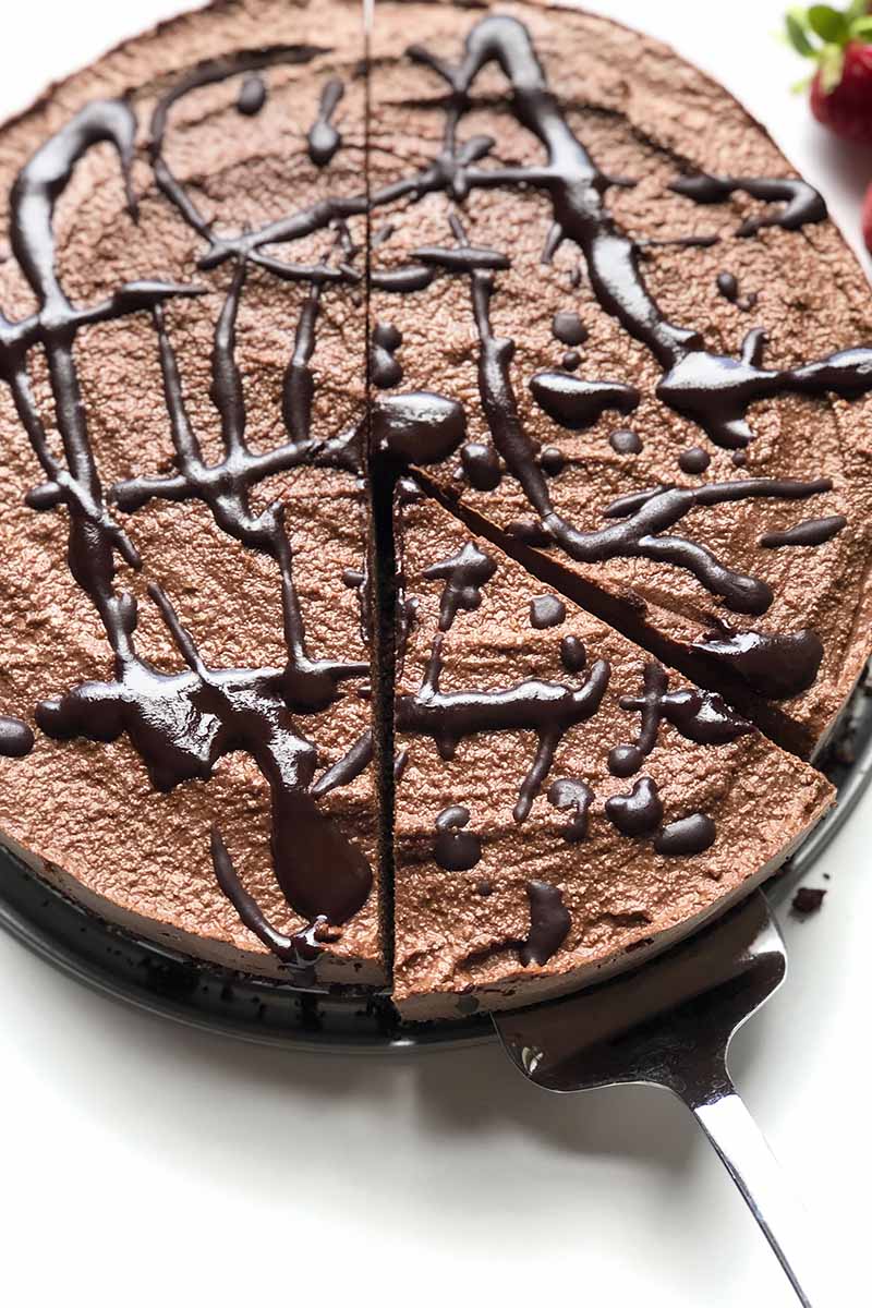 Vertical image of a whole chocolate cheesecake with dark drizzle, with a piece being removed from it with a metal serving knife.
