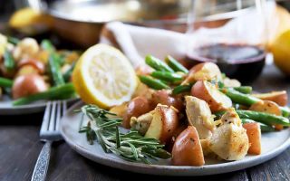 Horizontal image of two plates with potatoes, green beans, and cooked poultry pieces next to lemon wedges on a wooden table with a metal fork, towel, and glass of red wine.