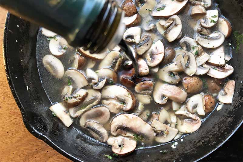 Horizontal image of added a dark liquid to a pan of sliced cooked mushrooms.