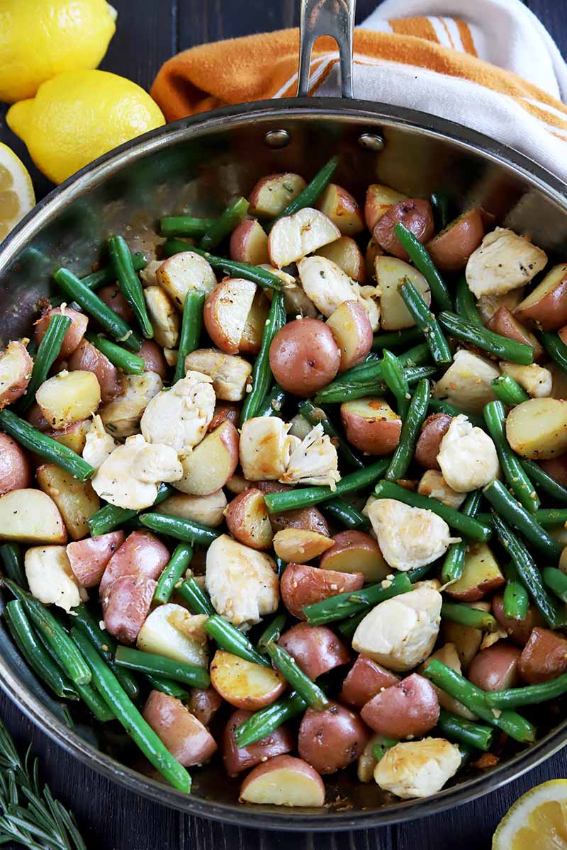Vertical top-down image of a skillet filled with potatoes, green beans, and cooked poultry pieces next to lemon wedges and a towel.