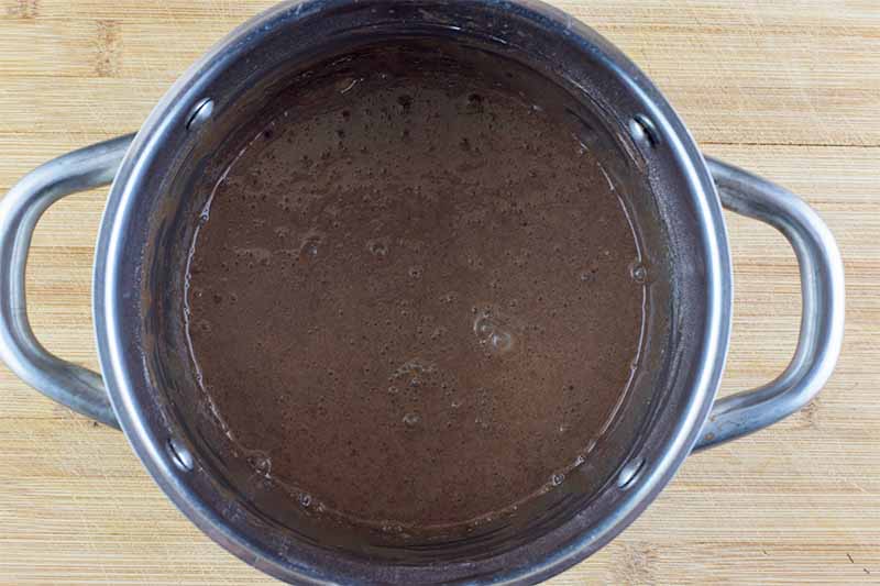 Horizontal image of a dark brown liquid in a metal pot on a wooden surface.