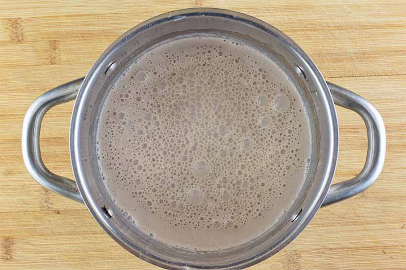Horizontal image of a frothy dark brown liquid in a pot on a wooden table.