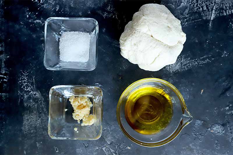 Horizontal image of a glass bowls of salt, garlic, and oil next to a mound of uncooked dough on a dark surface.