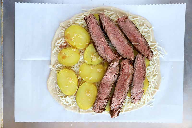 Horizontal image of slices of cooked meat, slices of spuds, and cheese on top of an unbaked crust on a parchment paper.