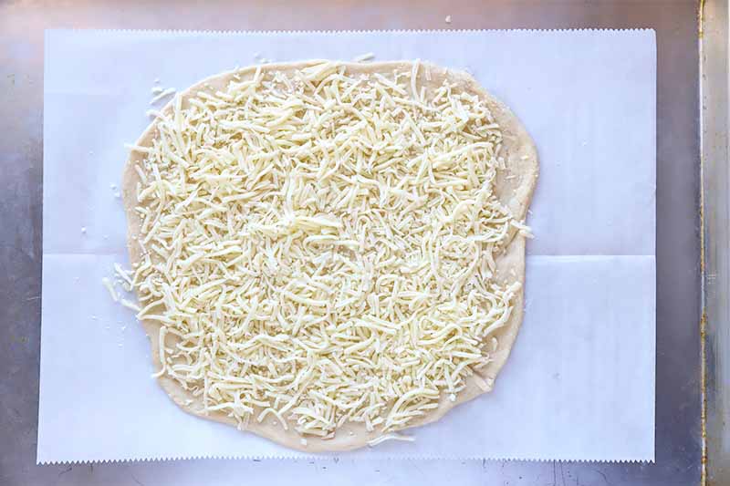 Horizontal image of shredded cheese on an unbaked dough crust on a parchment paper.