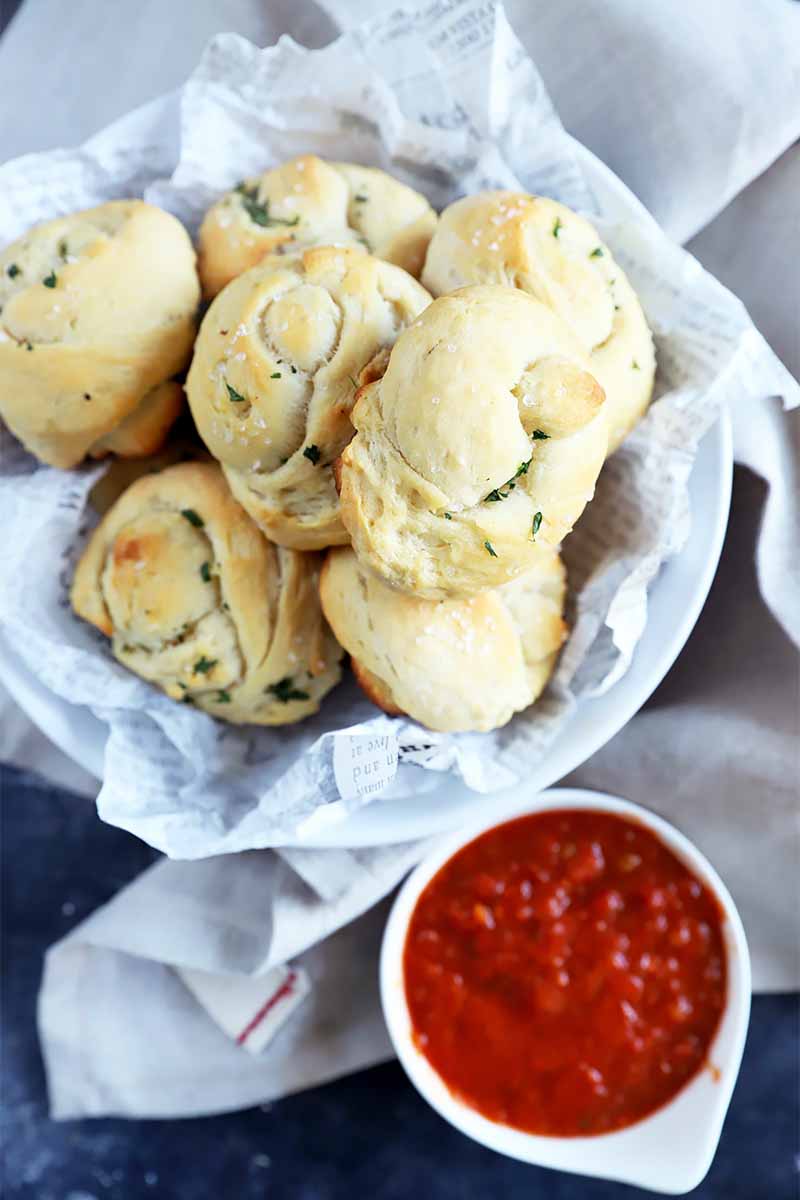 Vertical image of a pile of dinner rolls in a bowl next to marinara sauce in a white bowl.