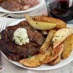 Horizontal image of a white plate with a seared large cut of beef topped with a dollop of herb butter next to potato wedges next to a glass of wine.